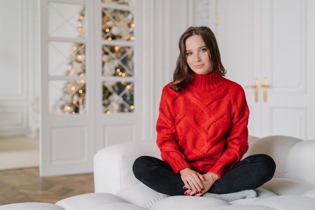 woman dressed in warm red knitted sweater, sits crossed legs in lotus pose on couch.