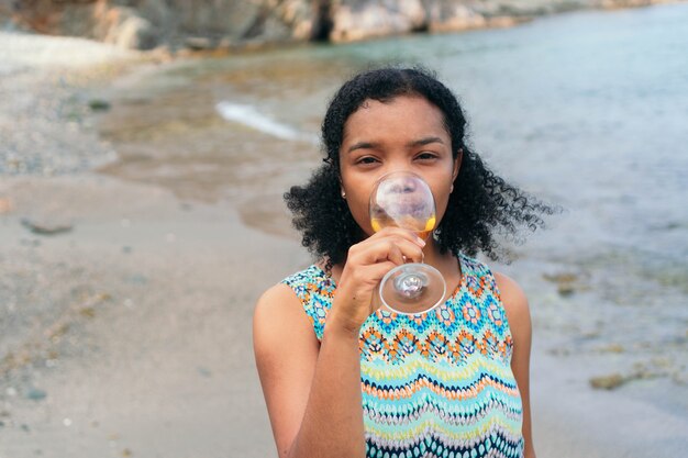 A woman drinking a glass of wine looking at the camera by the sea.