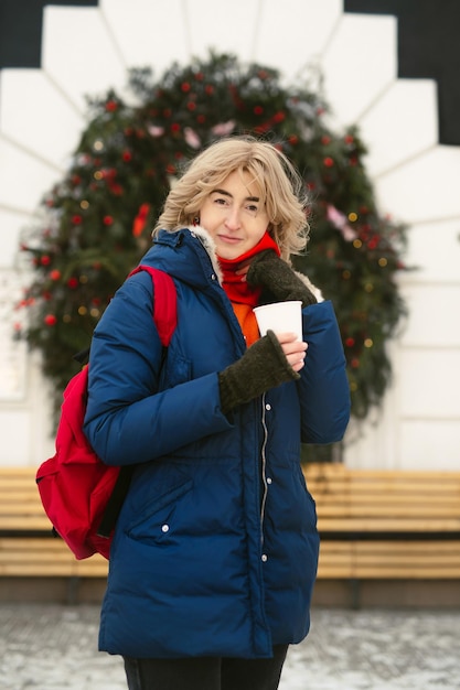 Woman drinks a hot drink on a city street against the background of a cafe window which is decorated