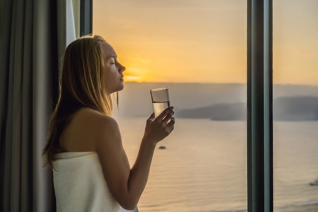 Woman drinks water in the morning on a background of a window with a sea view