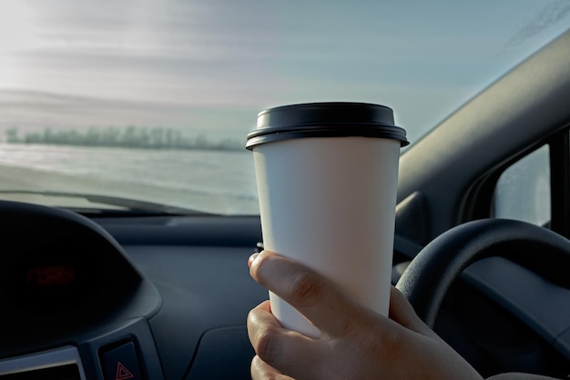 A woman drives a car and holds in her hand a white paper cup of coffee or a drink Layout for the inscription