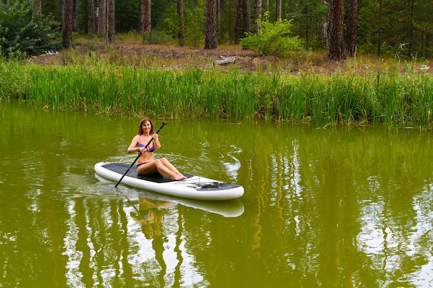 A woman drives on the Sup Board through a narrow canal surrounded by dense grass Active weekend vacations wild nature outdoor A woman is sitting with her legs stretched out