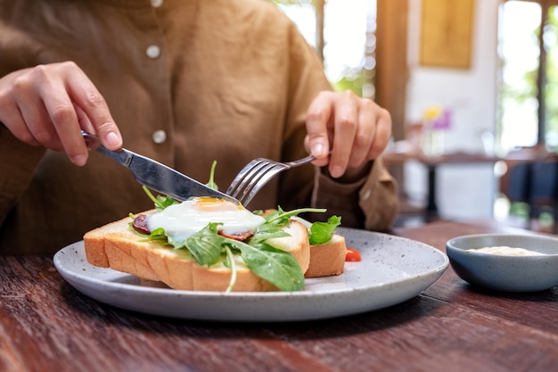 A woman eating breakfast sandwich with eggs, bacon and sour cream by knife and spoon in a plate on wooden table