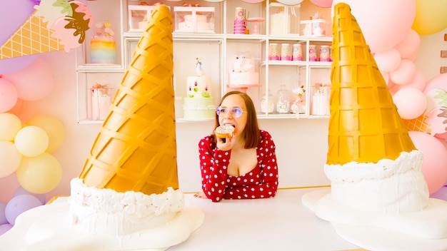 Woman eating doughnut near decorative ice creams Young female eating sweet doughnut at table with large decorative ice cream cones while working in light confectionery with balloons