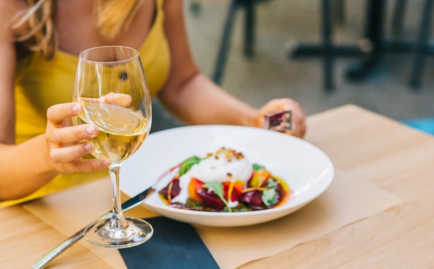 Woman eating healthy salad with burrata cheese, arugula salad and tomatoes and holding white wine in a glass