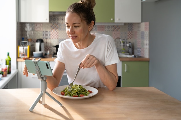 Woman eating pasta with vegetarian pesto sauce, addicted to social networks, watching movie on phone