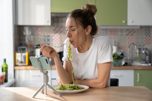 Woman eating pasta with vegetarian pesto sauce, watching movie on smart phone on tripod at home.