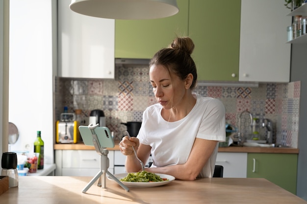 Woman eating pasta with vegetarian pesto sauce, watching movie on smart phone on tripod at home.