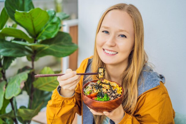 Woman eating raw organic poke bowl with rice and veggies closeup on the table top view from above