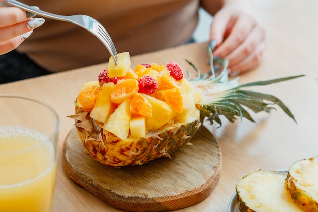 Woman eating tasty tropical fruit salad in half pineapple. Berries raspberries pineapple tangerines oranges in dessert fruit salad in cafe. Food fruit served in pineapple. High quality stock photo.