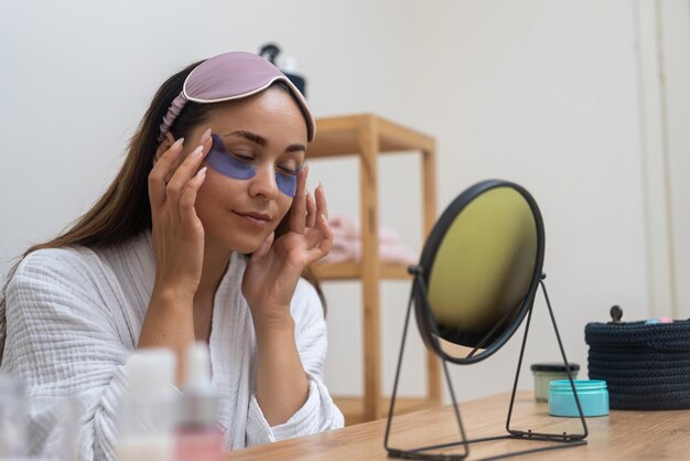 Woman enhances her antiaging skincare using eye patches and gazing to mirror during a home spa day