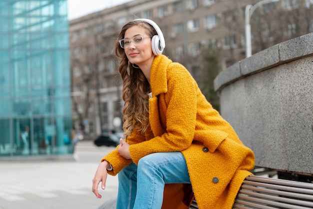 Woman Eyeglasses Listen Music Headphone Outdoor Sitting Bench Outdoor Dressed Stylish Yellow Coat Smile Caucasian Female 30s Enjoy Podcast Or Audio Books Outside