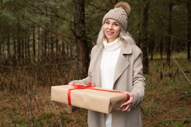 Woman in fashion warm clothes holding gift box in Kraft paper packaging on nature background