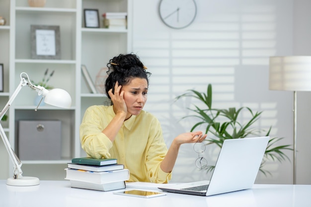 Woman feeling frustrated while working on a laptop in a home office setting with books and paperwork