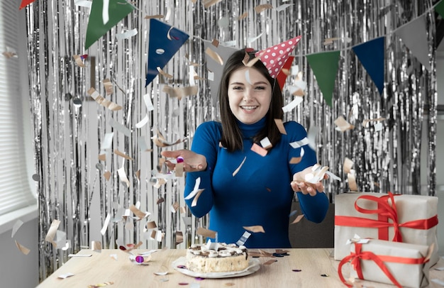 Woman in a festive cap sits at the table at home and celebrates her birthday confetti falls on her woman portrait look into the camera