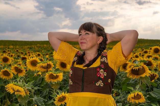 Woman in a field of sunflowers and she wears a yellow blouse a brown sundress and a yellow apron