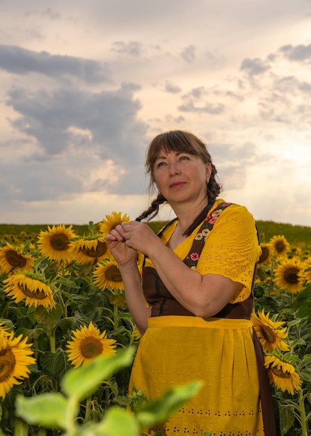 Woman in a field of sunflowers and she wears a yellow blouse a brown sundress and a yellow apron