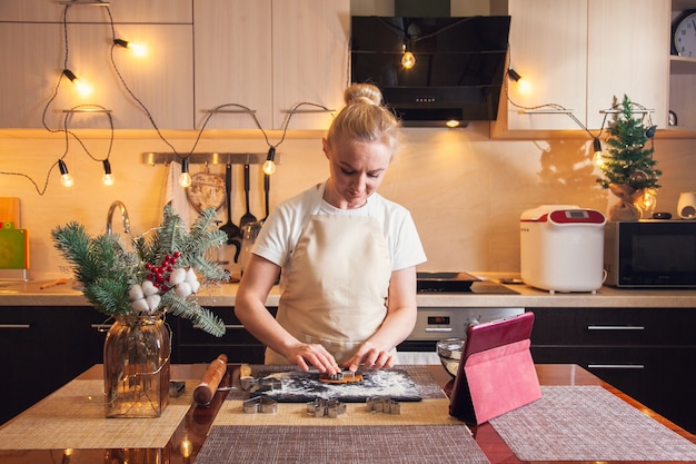 Woman following recipe on digital tablet and cuts out dough with mold for cooking christmas gingerbread cookies on her kitchen.