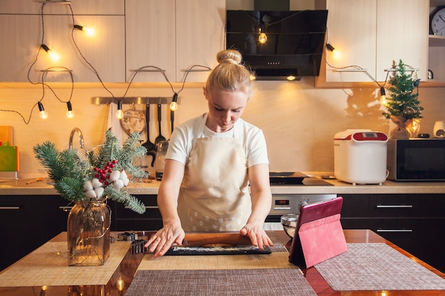 Woman following recipe on digital tablet and rolls out the dough with a rolling pin for cooking christmas gingerbread cookies on her kitchen.