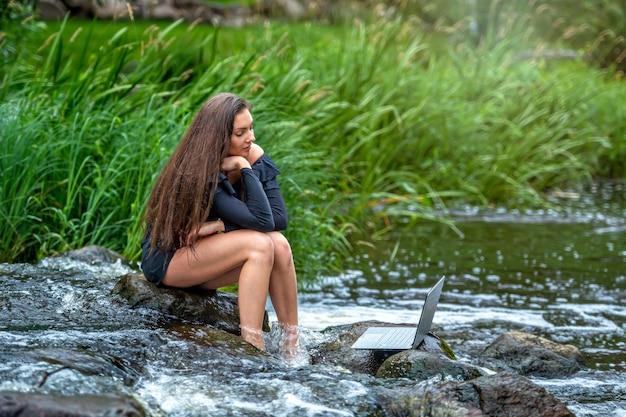 Woman freelancer sitting on a rock in the river and using a laptop travel or vacation work concept