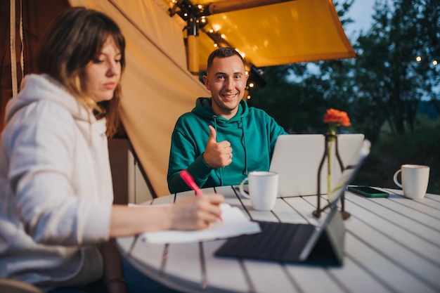 Woman freelancer working laptop and making notes while colleague looking camera sitting in cozy glamping tent in summer evening Luxury camping tent for outdoor holiday and vacation Lifestyle concept