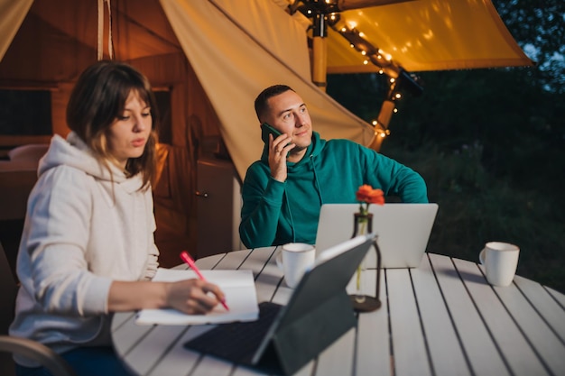 Woman freelancer working laptop and making notes while colleague talking phone sitting in cozy glamping tent in summer evening Luxury camping tent for outdoor holiday and vacation Lifestyle concept