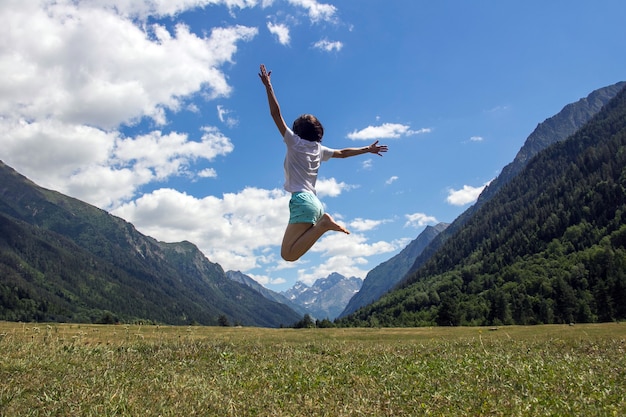 Woman full of joy jumps against mountains and a blue sky with white clouds Expression of love for travel and nature