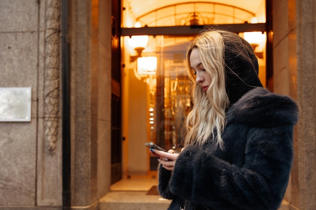 A woman in a fur coat texting on her phone outside a building