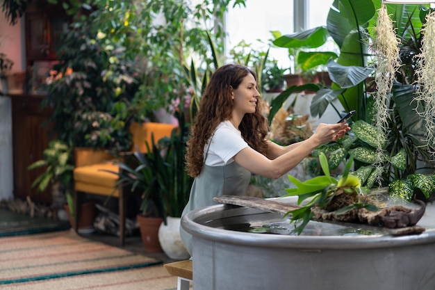 Woman gardener taking photo of aquatic plant in greenhouse using smartphone under freestanding bath