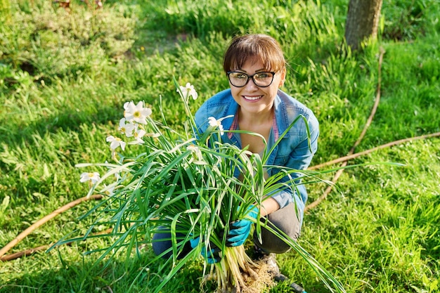 Woman in gardening gloves with white daffodil bush plant and shovel female looking at camera