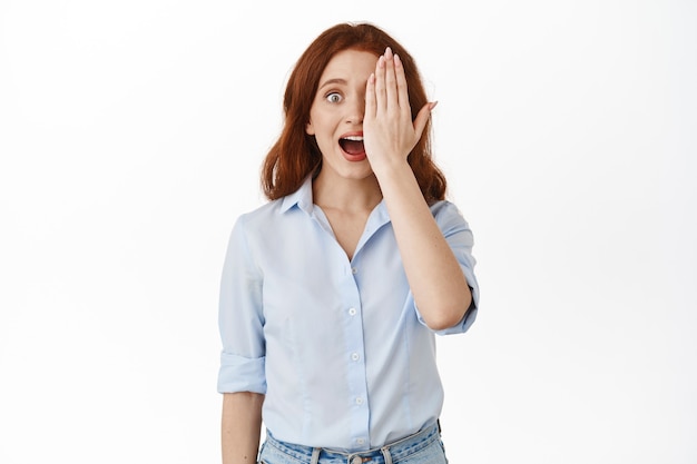 woman gasp from amazement, hide one half of face with palm, staring at something awesome, standing in blouse on white