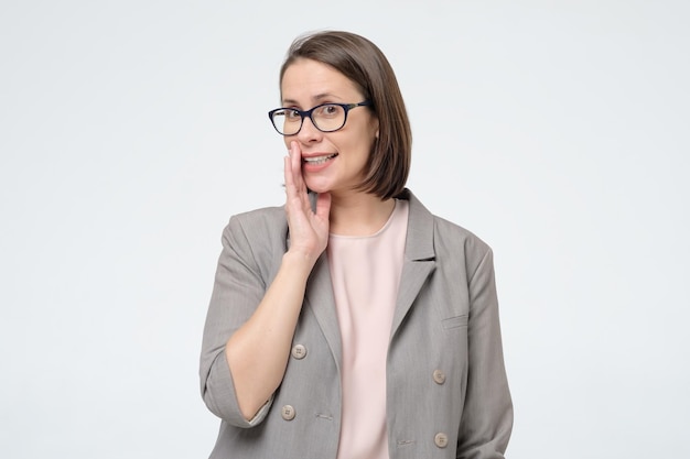 Woman in glasses and formal suit covering moth with hand tells secrets to someone