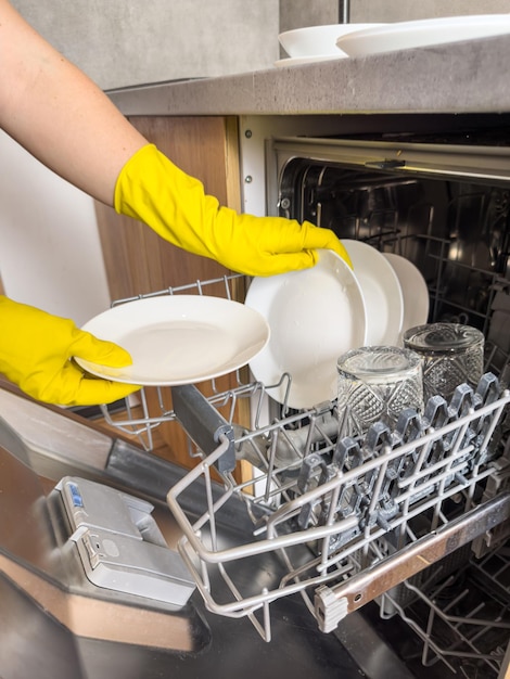 Woman in gloves placing plate and glasses in automatic builtin dishwashers machine