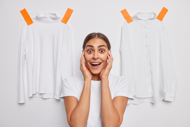 woman grabs face cannot believe her eyes stares wondered poses on white with plastered turtleneck and shirt on white studio wall