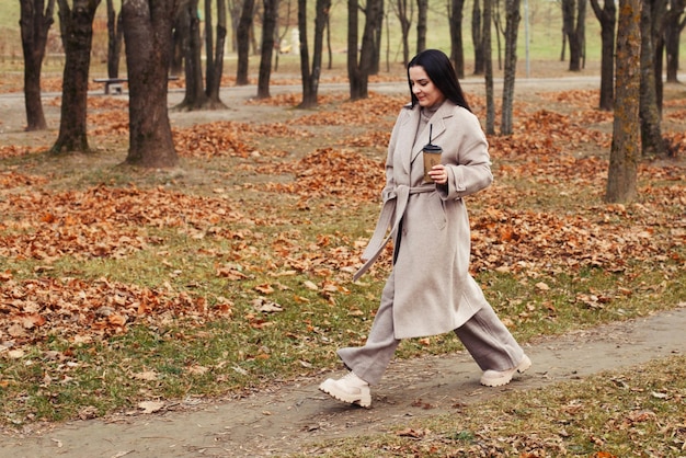 Woman in a gray coat walking in the autumn park with a cup of hot drink