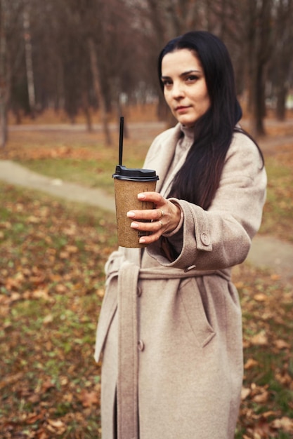 Woman in a gray coat walking in the autumn park with a cup of hot drink