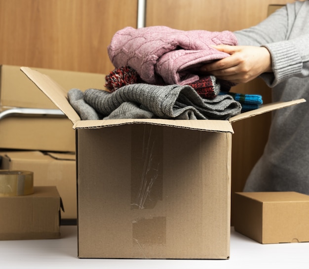 Woman in a gray sweater collects clothes in a box, concept of assistance and volunteering, moving. Behind a stack of brown boxes