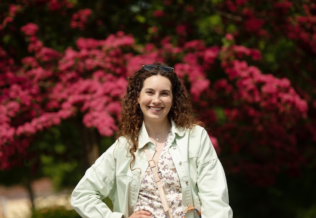 A woman in a green jacket is standing in front of a tree with pink flowers in the background.