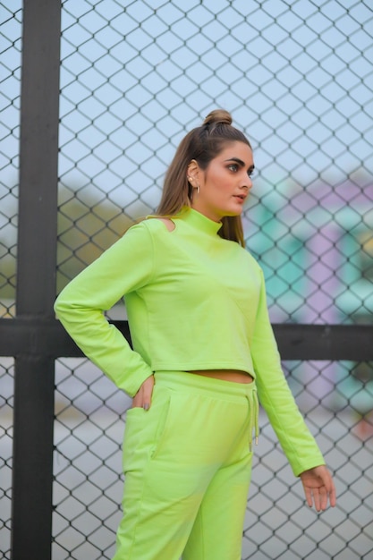 A woman in a green outfit stands in front of a fence wearing a green track suit.