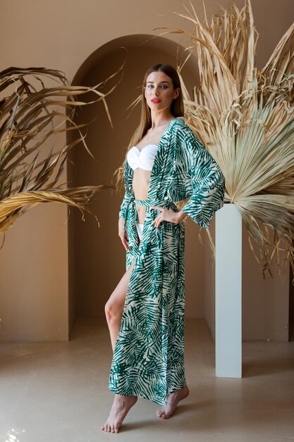 A woman in a green palm leaf kimono stands in front of a plant