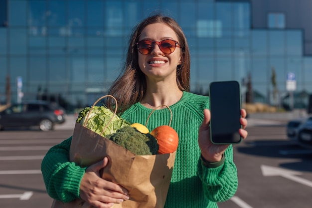 Woman in green sweater holding paper bag with full of groceries in hands