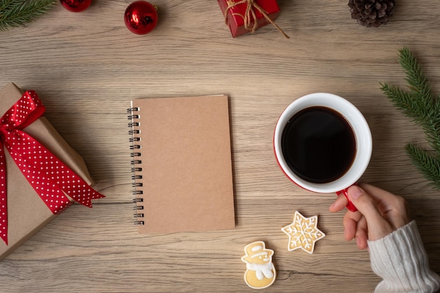 Woman hand holding black coffee cup with blank notebook and Christmas cookies on table. Xmas, Happy New Year, Goals, Resolution, To do list, Strategy and Plan concept