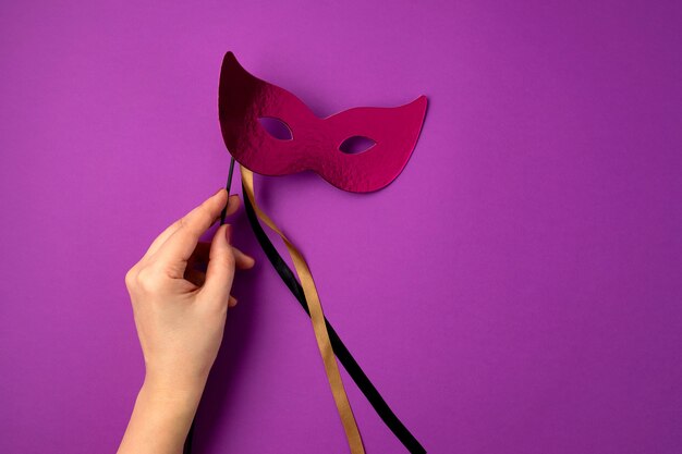 Woman hand holding  festive, colorful mardi gras or carnival mask over purple wall. Flat lay, top view