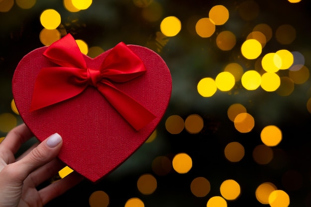 Woman hand holding a heart shaped red gift box with a bow on a blurry lights background. Empty space