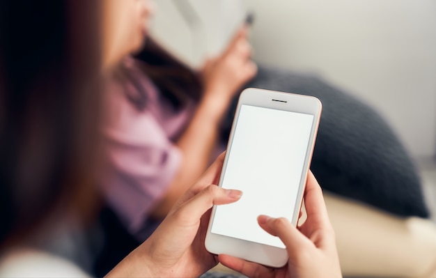 Woman hand holding smartphone with blank screen on sofa in house with friends. Take your screen to put on advertising.