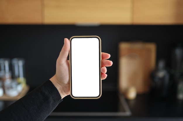 Woman hand holding smartphone with vertical white screen on kitchen at home