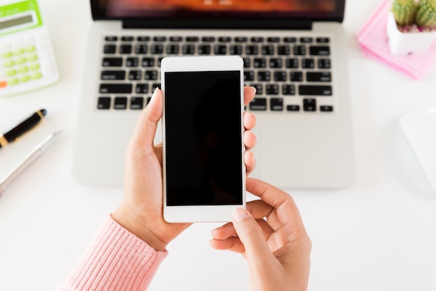 Woman hand holding white mobile phone on a table with a laptop in office