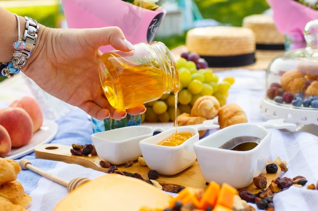 Woman hand pours honey from transparent bottle to sauceboat, at summer picnic background