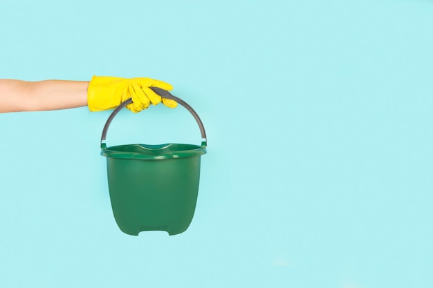 Woman hand with yellow rubber glove holding a green pail on a light blue background with copy space