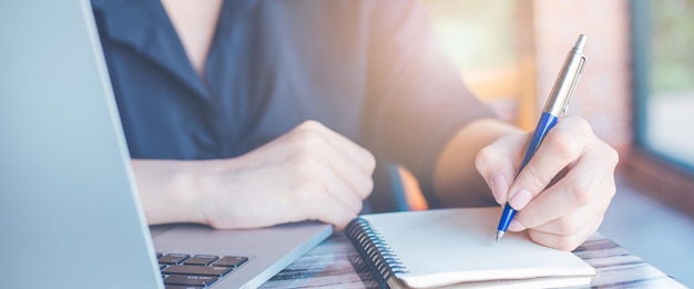 Woman hand writing on a notepad with a pen and use a laptop computer in the office.for web banner.